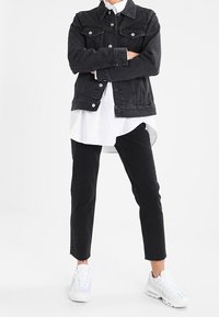 Czarna kurtka dżinsowa z dwoma kieszeniami na piersi, nałożona na białą koszulę, zestawiona z czarnymi, podwiniętymi jeansami i białymi trampkami.