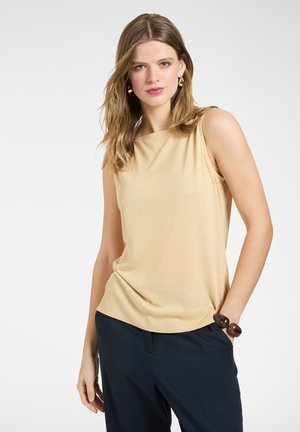 Kobieta ubrana w beżowy top bez rękawów, granatowe spodnie, duża ciemna bransoletka i złote kolczyki, pozująca na tle jasnego, jednolitego tła.