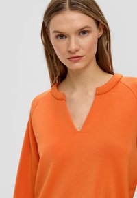 Oranje pullover met een V-hals en ronde kraag. Gemaakt van zachte, gestructureerde stof met raglanmouwen. Eenvoudig en modern ontwerp.