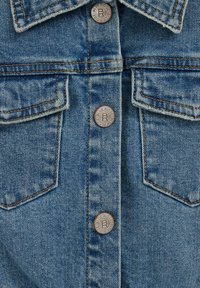 Denim jaka ar vara krāsas metāla pogām, kontrastējošām šuvēm un divām krūšu kabatām. Gaiši zila auduma ar teksturētu apdari.