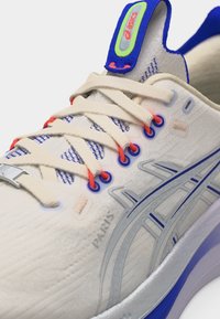 Prim-plan cu o pantof sport ASICS alb, cu șireturi bej, orificii roșii, accente albastre și cuvântul „Paris” gravat pe lateral.