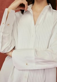 Chemise blanche boutonnée avec plis verticaux, ornée de boutons de manchette en or, larges poignets et col classique. Texture lisse et nette.