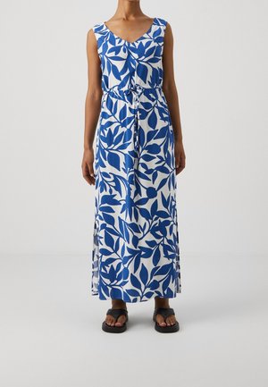 Ärmelloses weißes Maxikleid mit blauem Blattmuster, tailliert, getragen mit schwarzen flachen Sandalen an einer Person, die vor einem einfarbigen Hintergrund steht.