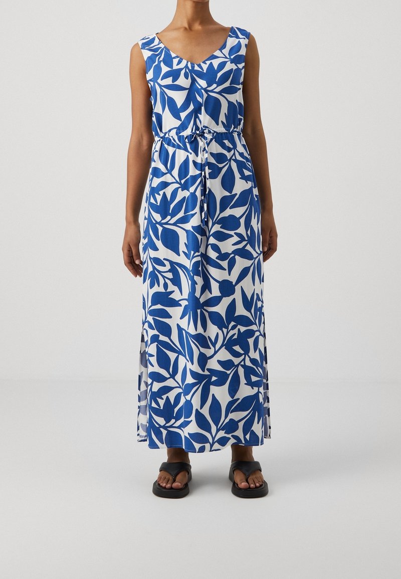 Robe maxi blanche sans manches avec imprimé feuilles bleues, taille cintrée, portée avec des sandales plates noires sur une personne debout devant un fond uni.