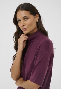 Oversized, short-sleeve top in deep purple fabric with a high collar. Soft texture, minimal stitching details. Model poses thoughtfully.
