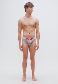 Sive bombažne hlačke z rožnatim pasom, na katerem je rdeče napisano "SEX". Model stoji obrnjena naprej, bosa, brez zgornjega dela oblačila.