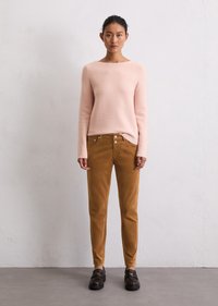 Jasnoróżowy sweter z długim rękawem, zestawiony z brązowymi spodniami z korduroyu z przednimi kieszeniami i zapięciem na guzik. Ciemne buty dopełniają look.