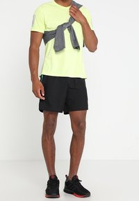 Camiseta atlética de manga corta amarilla brillante, pantalones cortos negros, sudadera gris atada a la cintura, zapatillas negras con acentos rojos.
