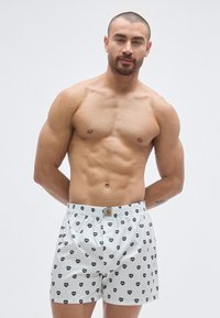 Boxer bianchi con motivo ripetuto di cuori neri, vita elastica e etichetta sul davanti. Modello a torso nudo che presenta il prodotto.