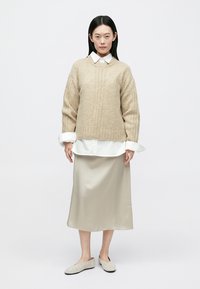 Μπεζ πλεκτό πουλόβερ με ριμπ μοτίβο, φορεμένο πάνω από άσπρο πουκάμισο με γιακά, συνδυασμένο με μπεζ σατέν φούστα και ανοιχτά παντόφλες.