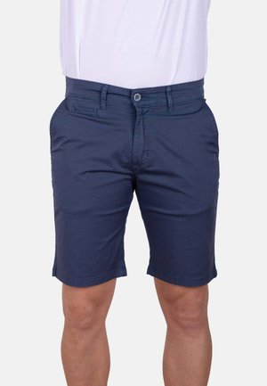 Hombre vestido con pantalones cortos azul marino hasta la rodilla y una camisa blanca, mostrado de la cintura a las rodillas sobre un fondo liso.