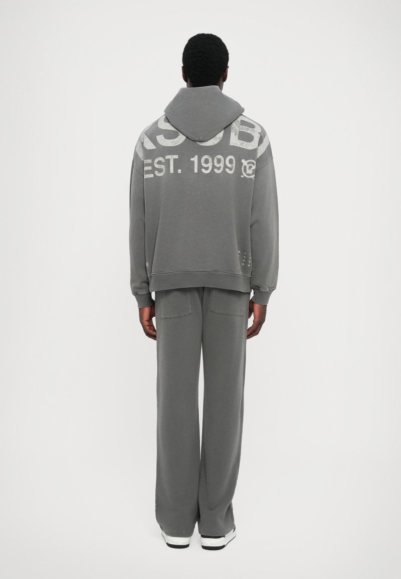 Gray sweatshirt and pants set with a relaxed fit. Sweatshirt has a hood and large white graphic text on the back. Soft fabric texture.