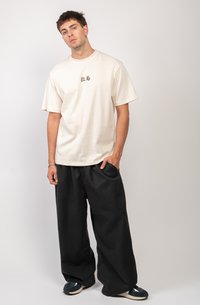 Camiseta color crema de manga corta con texto negro en el pecho, combinada con pantalones negros de corte holgado y zapatillas deportivas oscuras.