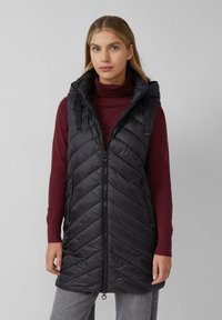Schwarze Pufferweste mit Chevron-Muster, Reißverschluss vorne und Kapuze. Getragen über einemburgunderfarbenen Rollkragenpullover und mit zwei Seitentaschen.