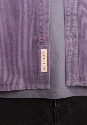 Paarse corduroy shirt met een knoopsluiting en een label dat "REVOLUTION" zegt. Draagt over een grijs T-shirt en zwarte broek.