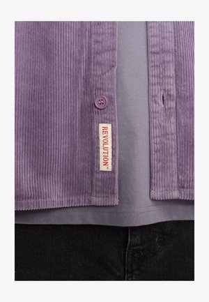 Camicia di velluto a coste viola con chiusura a bottoni e un'etichetta che recita "REVOLUTION". Indossata sopra una maglietta grigia e pantaloni neri.