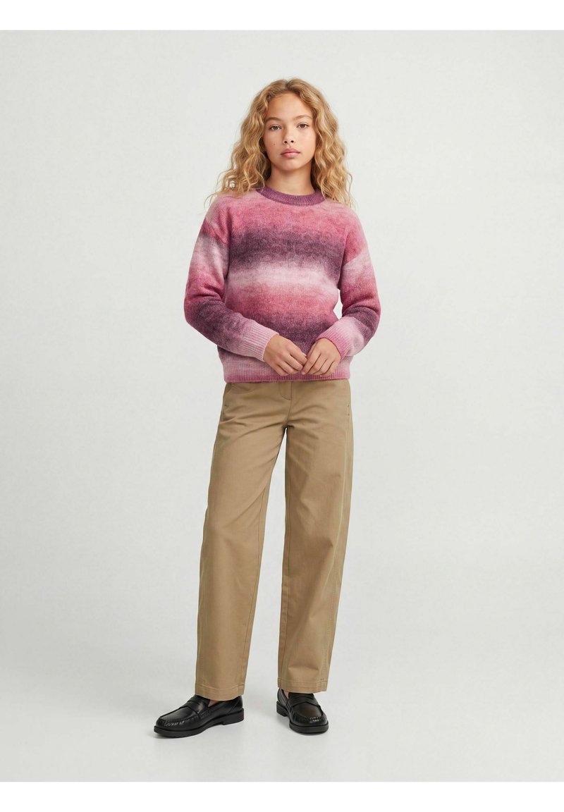 Jeune personne debout portant un pull rayé rose et violet, un pantalon beige et des chaussures noires sur un fond clair uni.