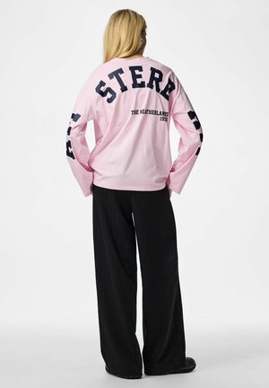 Personne aux longs cheveux blonds portant un haut rose à manches longues avec les inscriptions "STER" et "The Netherlands 1993", ainsi qu'un pantalon large à rayures noires.