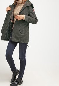 Parka verde con cappuccio foderato in pelliccia, chiusura a boton e zip, due tasche frontali e una toppa sulla manica. Indossato sopra un maglione beige e jeans scuri.