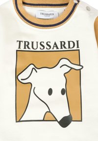 Felpa bianca con maniche beige. Presenta una grafica della testa di un cane in contorno nero su uno sfondo quadrato giallo senape. Logo "TRUSSARDI" sopra.