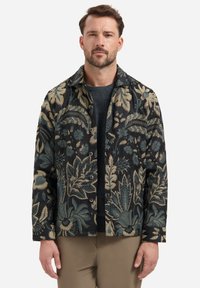 Hemdjack met bloemenpatroon in donkere stof met blauwe en beige ontwerpen, met knoopsluitingen en een kraag. G gedragen over een grijze trui.