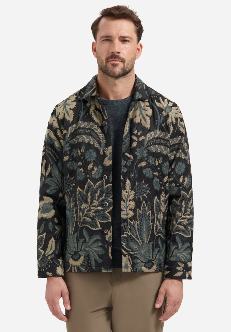 Hemdjack met bloemenpatroon in donkere stof met blauwe en beige ontwerpen, met knoopsluitingen en een kraag. G gedragen over een grijze trui.