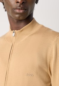 Close-up van een persoon die een beige ritsvest draagt met een geribbelde kraag en 'BOSS' geborduurd op de borst.