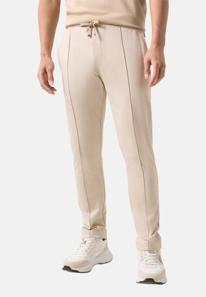 Mężczyzna ubrany w beżowe, dopasowane spodnie o przednich szwach, z wiązanym paskiem w talii, w połączeniu z białymi i beżowymi trampkami, stojący na neutralnym tle.