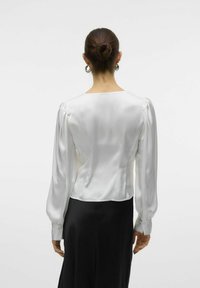 Blusa de satén blanca con mangas largas abullonadas, un escote cuadrado y puños abotonados. Llevada con una falda negra, que muestra una textura suave.