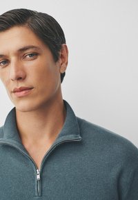 Pilkas zip-up megztinis su aukštu koloriumi, pagamintas iš minkštos mezginio medžiagos. Įrengtas sidabriniu užtrauktuku ir lygiu paviršiumi. Detalus vaizdas ties iškirpte.