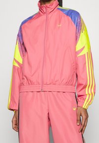 Osoba oblečená v růžové teplákové soupravě Adidas s žlutými a fialovými detaily na rukávech a žlutými logy Adidas na hrudi a kalhotách.