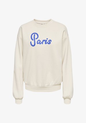 Vrouw draagt beige sweater met opschrift "Paris", blauwe spijkerbroek en lichtblauwe hakken, houdt witte kanten stof diagonaal over haar heupen.