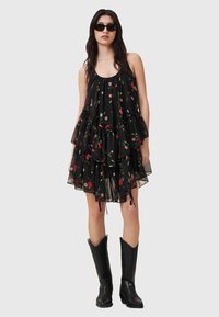 Schwarzes Blumenkleid mit mehreren Rüschenschichten, mit roten und grünen Mustern. Getragen mit schwarzen Sonnenbrillen und knielangen schwarzen Cowboy-Stiefeln.