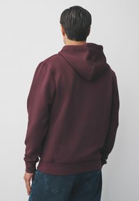 Burgunderfarbener Kapuzenpullover mit strukturiertem Muster, eng anliegenden Bündchen und einer Kängurutasche, kombiniert mit dunklen Jeans.