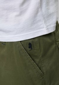 Camiseta blanca de algodón superpuesta sobre pantalones verde oliva. Los pantalones cuentan con un bolsillo lateral y un pequeño detalle de logo bordado en negro.
