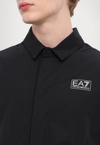 Svart lättviktsjacka med knapplik krage, med vertikala sömmar och en EA7 Emporio Armani-logotyp på bröstet.