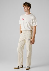 Crème t-shirt met rood logo, gecombineerd met lichtbeige cargobroek met utility pockets en een stoffen patch. Bruin en witte sneakers.