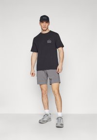 T-shirt en coton noir avec motif de poche, shorts gris en matériau léger, chaussures de sport grises avec lacets, et une casquette noire.