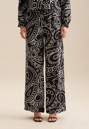 Schwarzfarbige Hochwasserhosen aus Stoff mit einem auffälligen weißen Paisleymuster. Elasthanbund, glatte Textur und offene Säume. Schwarze Sandalen.