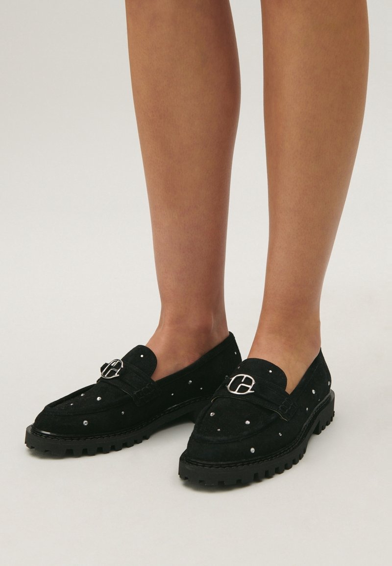 Mocassins en daim noir avec clous argentés et détail de boucle métallique, portés pieds nus, debout sur un fond clair uni.