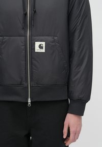 Schwarze, gepolsterte Bomberjacke mit gerippten Bündchen und Saum; verfügt über einen Reißverschluss auf der Vorderseite und ein quadratisches Logo-Patch auf der linken Brust.