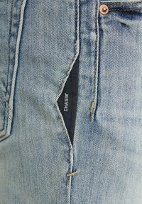 Jeans de mezclilla azul claro con un detalle decorativo de corte en el bolsillo. Presentan un respaldo de tela negra en contraste con la etiqueta de la marca "CHASIN'".