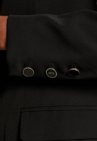 Puño de tela negra con tres botones redondos. Dos son completamente negros; uno tiene un hardware dorado texturizado con el texto "ICON" visible.