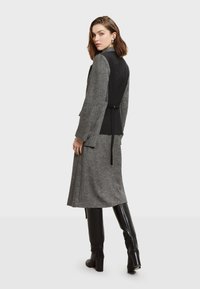 Dlhé šedé šaty z vlnenej zmesi s čiernym priliehavým korzetom a opaskom v páse. Majú dlhé rukávy a jemne textúrovaný povrch, kombinované s čiernymi čižmami po kolená.