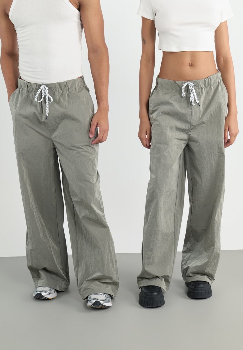 Deux personnes portant des pantalons gris larges à cordon, des hauts blancs et des baskets, debout côte à côte devant un fond uni.