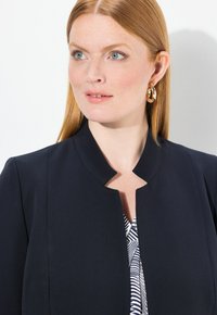 Blazer blu navy con scollo rotondo, realizzato in tessuto testurizzato. Presenta una chiusura nascosta e indossato sopra una camicia a fantasia.