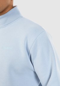 Sweatshirt bleu clair avec un col haut, fabriqué en tissu doux. Présente un logo brodé sur la poitrine dans une couleur assortie.