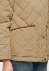 Μπεζ ριγωτό μπουφάν με σχέδια διαμαντιού, καφέ λεπτομέρειες στο τσεπάκι και κούμπωμα με πατιλέτα κουμπιών και λογότυπα.