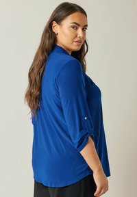 Blusa blu realizzata in tessuto morbido, con colletto, maniche arrotolabili e una vestibilità comoda con orlo curvo.