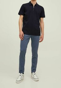 Hombre con una camiseta polo negra de manga corta, pantalones azul ajustados y zapatillas blancas, de pie contra un fondo claro y liso.